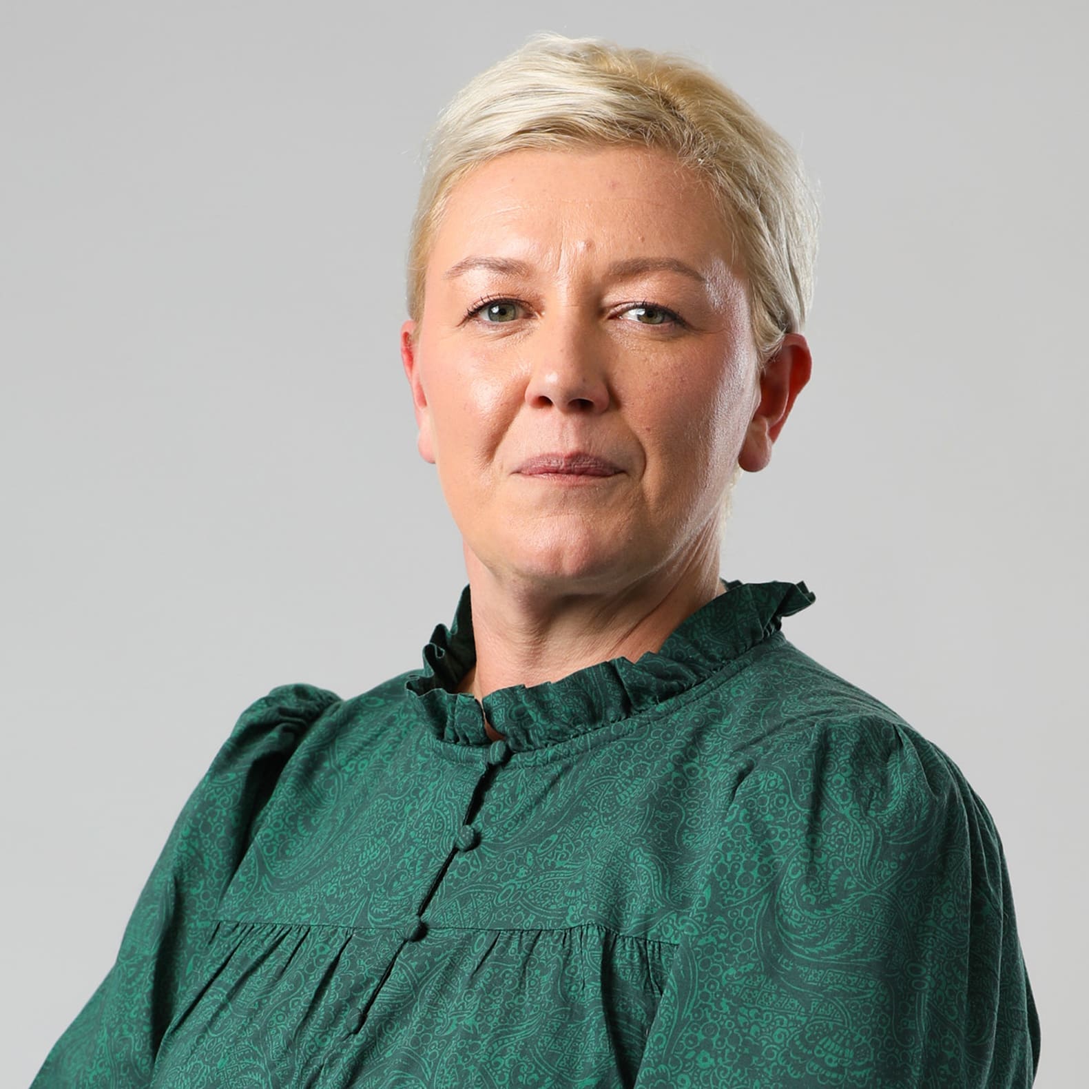 A woman with short blonde hair wears a green patterned blouse, looking confidently at the camera.